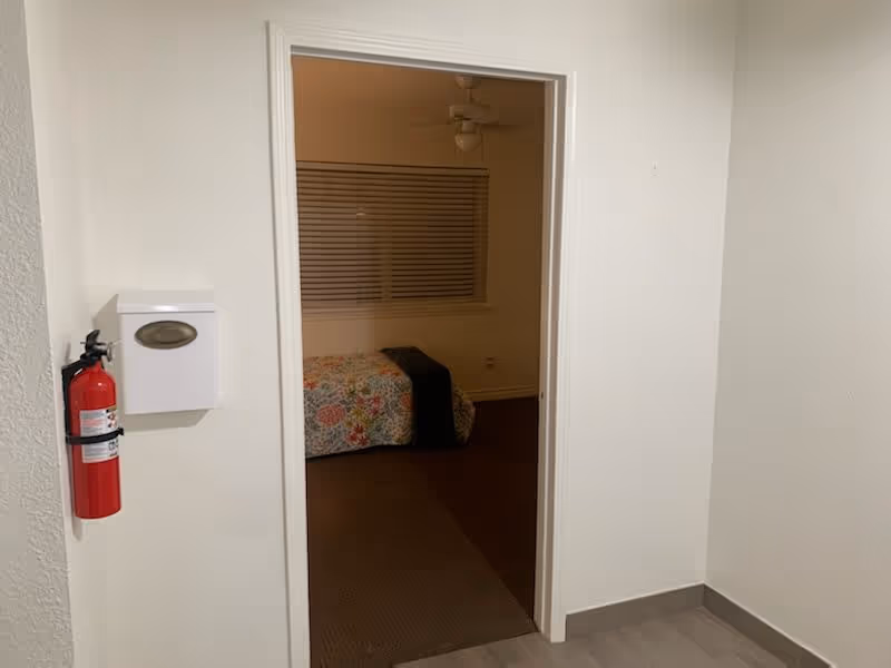 View through an open doorway into a bedroom with a bed covered in a colorful floral quilt and a dark blanket. The bedroom has a window with closed blinds and a ceiling fan. Outside the doorway, a fire extinguisher and a white wall-mounted box are visible on the left wall.