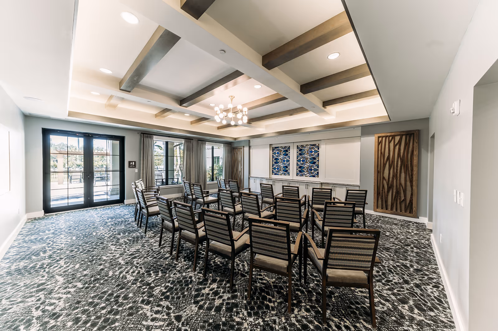 A bright multipurpose meeting room with rows of chairs, a coffered ceiling, patterned carpet, and glass doors to an outdoor area.