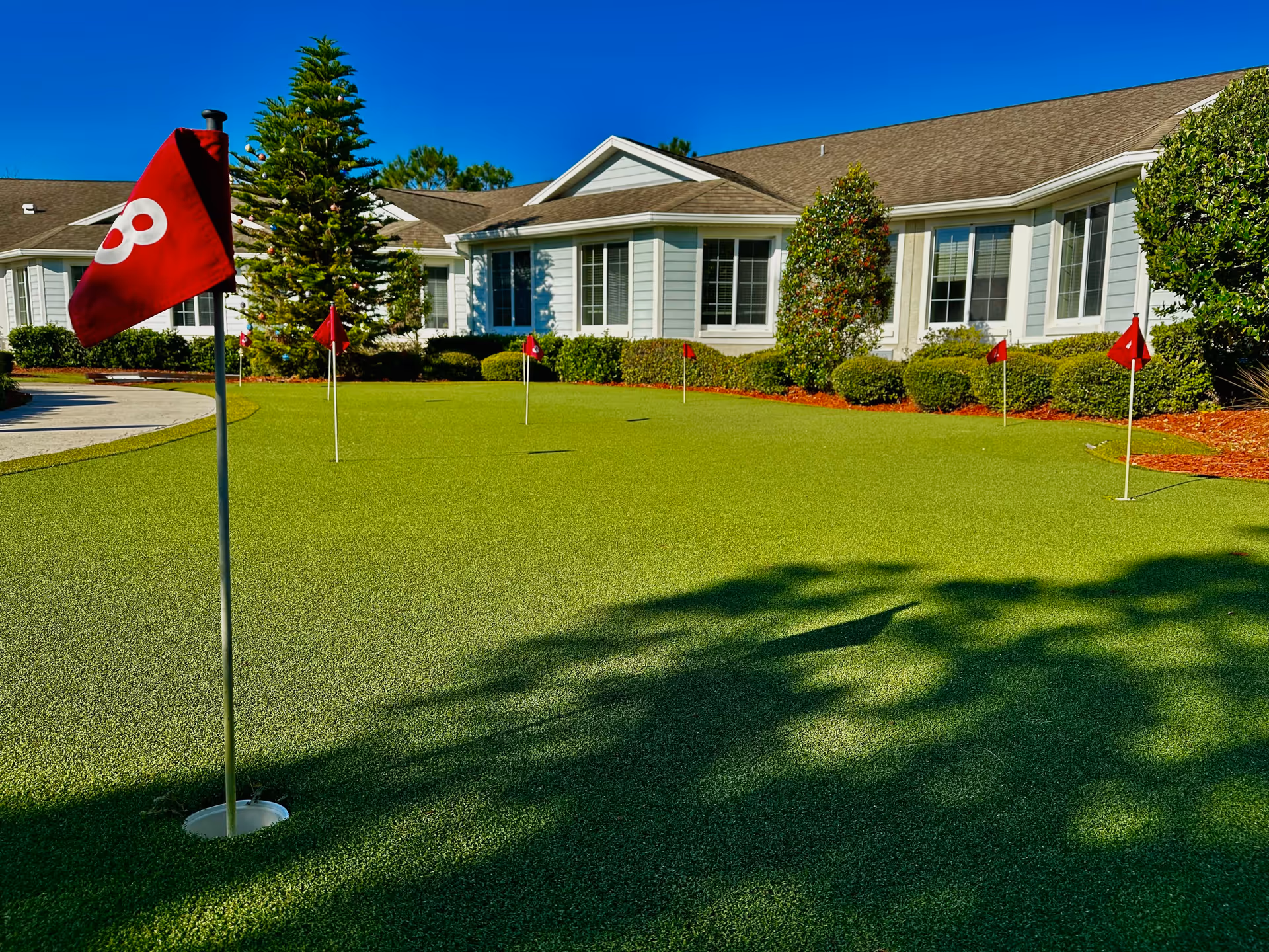 A putting green with several red flags and holes in front of a single-story building with white siding and multiple windows, surrounded by bushes and trees under a clear blue sky.