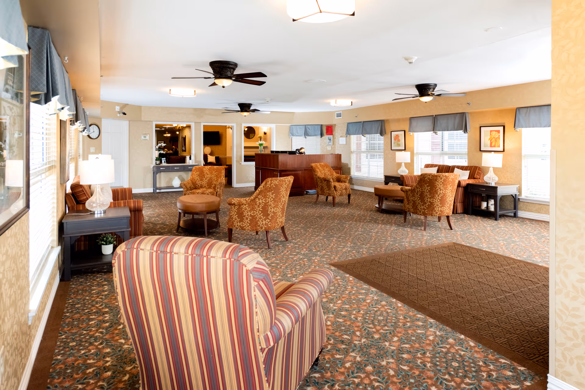 A spacious senior living facility common area with patterned carpet and multiple seating arrangements including striped and floral upholstered armchairs, ottomans, and side tables with lamps. The room has several windows with blue valances, ceiling fans, framed artwork on the walls, and a reception desk in the background.