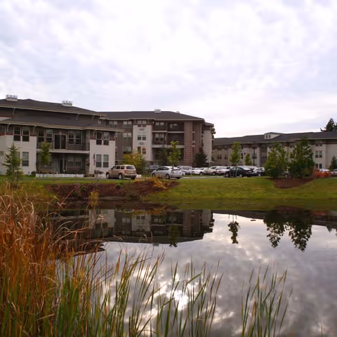 Exterior view of a multi-story senior living building with parked cars and a reflecting pond in the foreground.