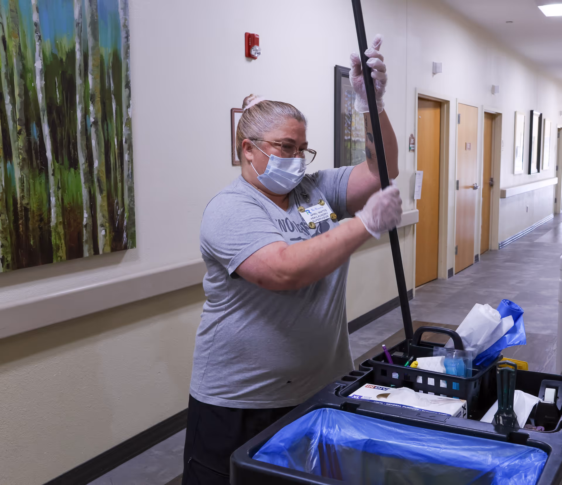 A woman wearing a face mask and gloves is cleaning a hallway in a skilled nursing facility. She is holding a mop handle and standing next to a cleaning cart filled with supplies. The hallway has several closed doors and framed artwork on the walls.