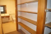 View of a bathroom through an open doorway from a small room with wooden shelves on the right side. The bathroom contains a toilet and a sink with a mirror above it. The walls are light-colored and the floor is beige.