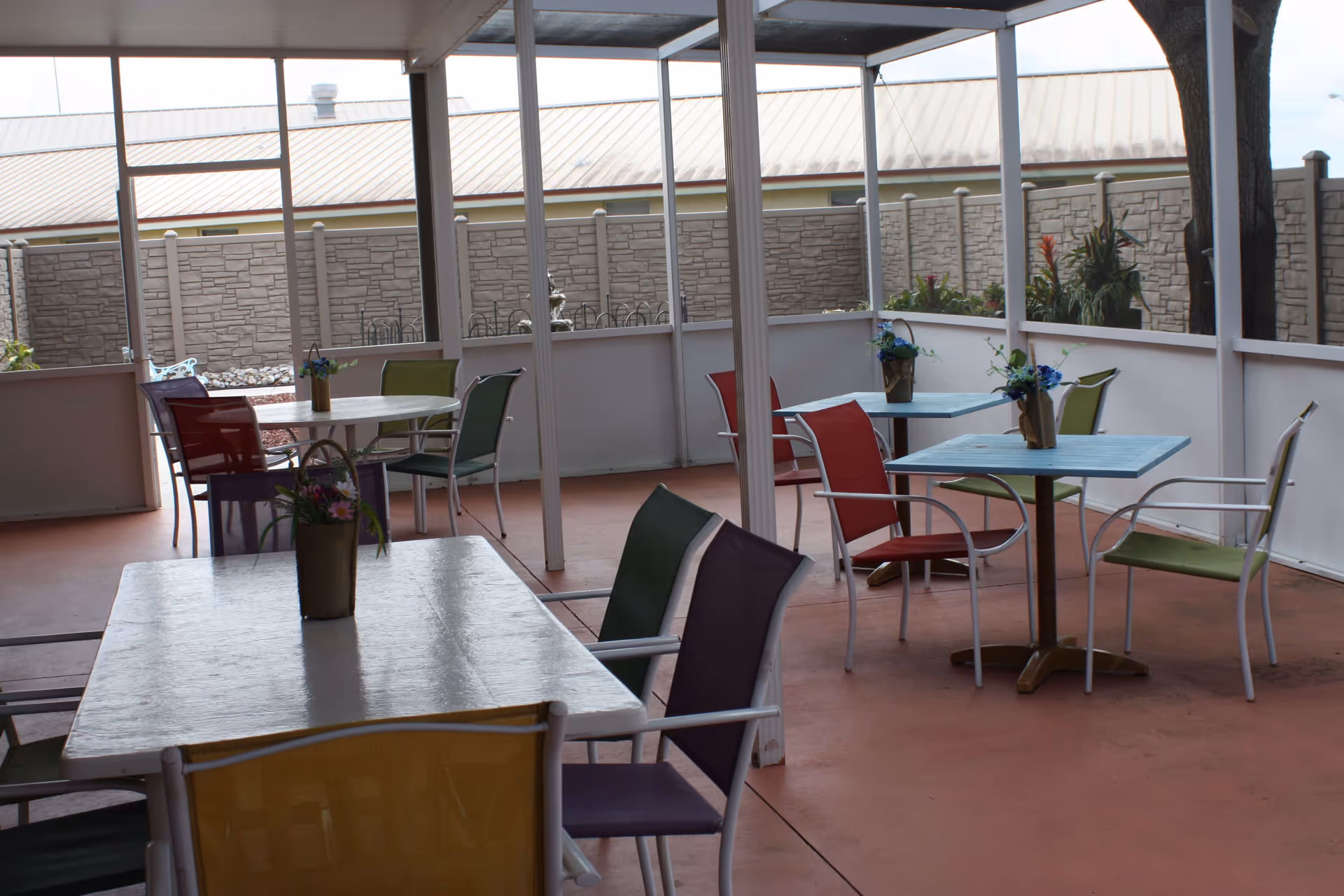 Covered outdoor patio area with several tables and colorful chairs arranged around them. Each table has a small flower arrangement in a vase. The patio is enclosed with white railings and has a stone wall and building visible in the background.