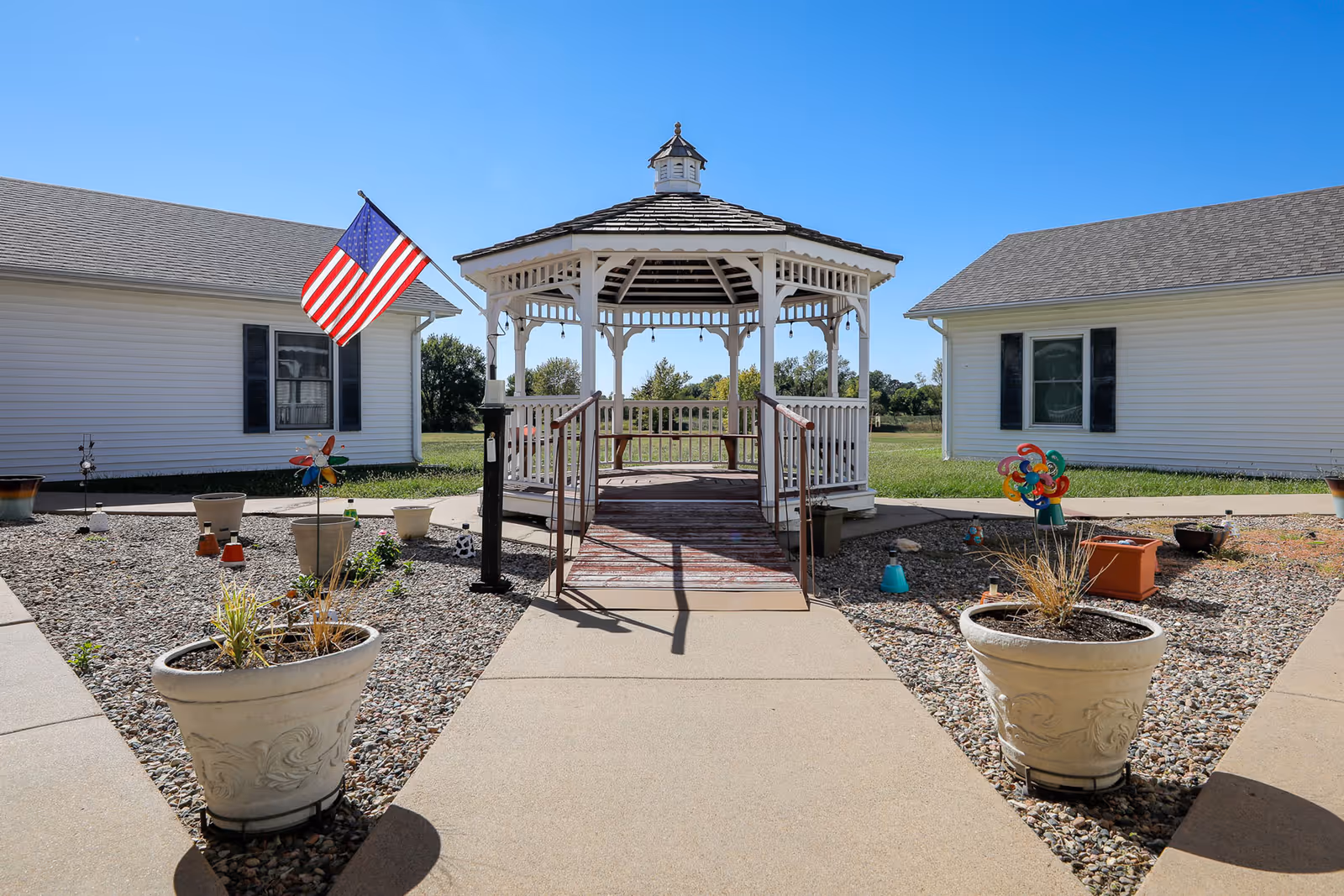Outdoor view of a white gazebo with a ramp entrance situated between two white buildings under a clear blue sky. There are potted plants and decorative items, including an American flag and colorful pinwheels, placed on a gravel area surrounding the walkway leading to the gazebo.