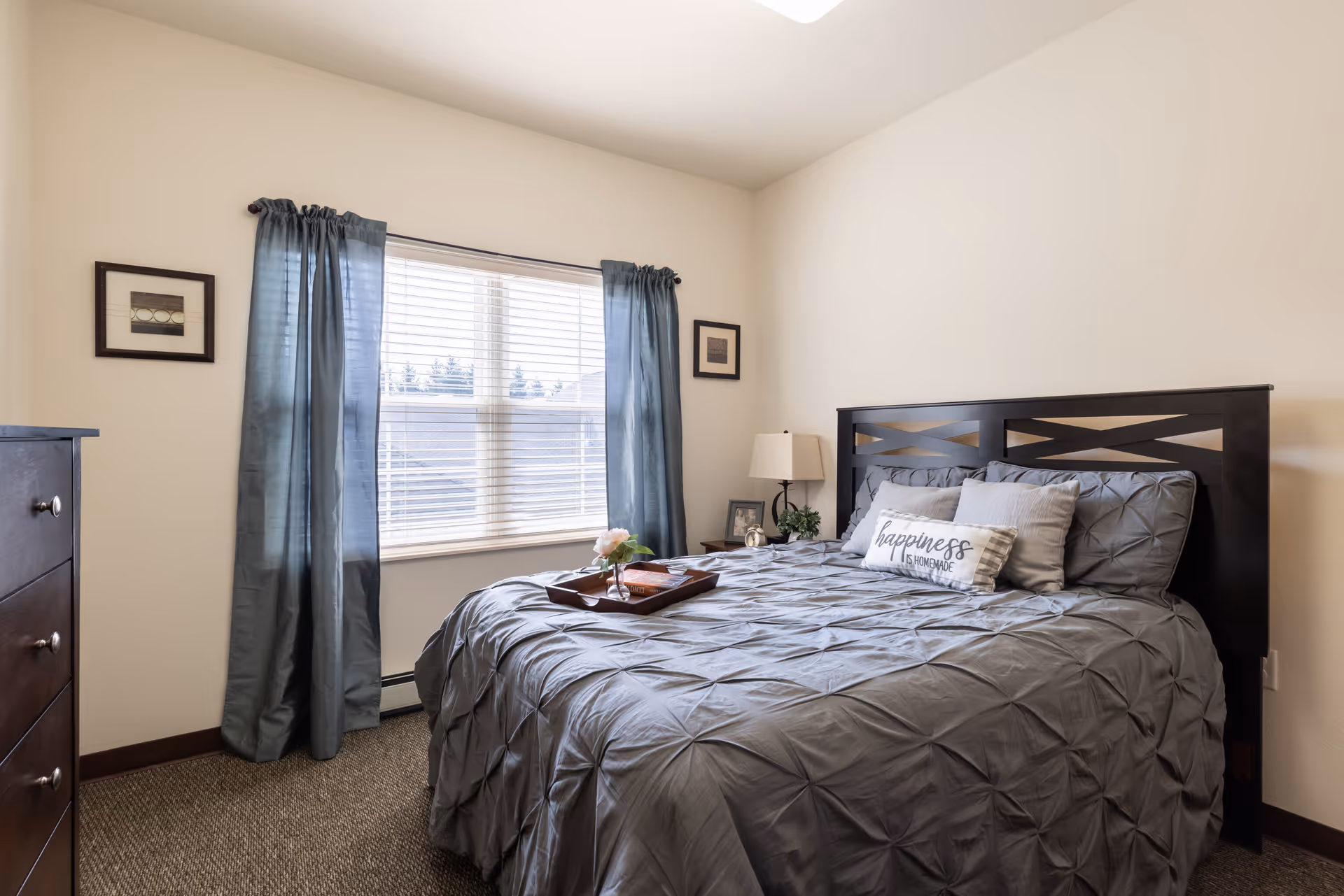 A cozy bedroom with a large bed covered in a gray comforter and multiple pillows, including one with the text 'happiness is homemade'. The room has a window with blue curtains, a wooden dresser on the left, a nightstand with a lamp and a small plant, and two framed pictures on the wall.