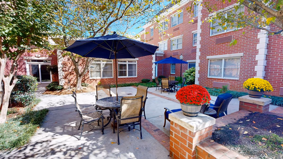 Outdoor courtyard area at Dunwoody Place featuring patio tables with umbrellas, chairs, potted red and yellow flowers, surrounded by brick buildings and trees with sunlight casting shadows.