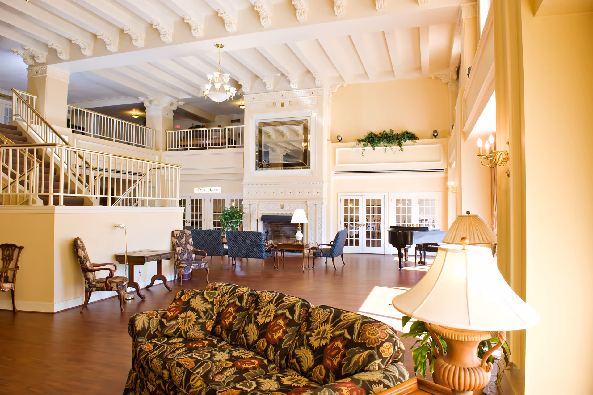 A spacious and elegant senior living community common area with high ceilings, decorative beams, and a chandelier. The room features a floral patterned sofa in the foreground, several upholstered chairs around a wooden coffee table near a large ornate fireplace, and a grand piano near glass-paneled double doors. There are stairs leading to an upper level with railings, and the room is warmly lit with lamps and wall sconces.