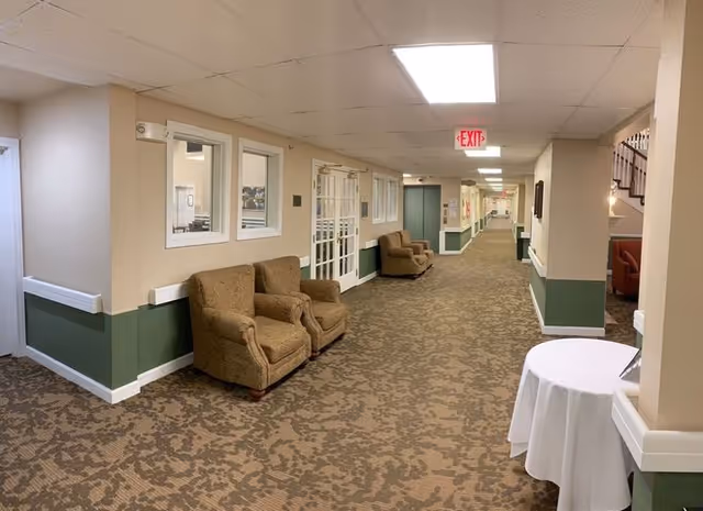 Long carpeted interior hallway with upholstered chairs along the wall, a small round table with a white cloth, and an EXIT sign overhead.