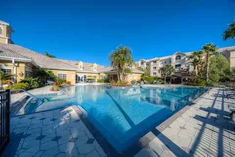 Outdoor swimming pool area at Aston Gardens At Pelican Pointe with clear blue water, surrounded by a tiled deck, palm trees, and multi-story residential buildings under a clear blue sky.