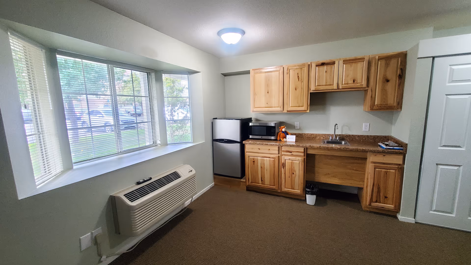 Small studio-style kitchenette with wooden cabinets, a mini fridge and microwave next to a large bay window and wall air conditioning unit.
