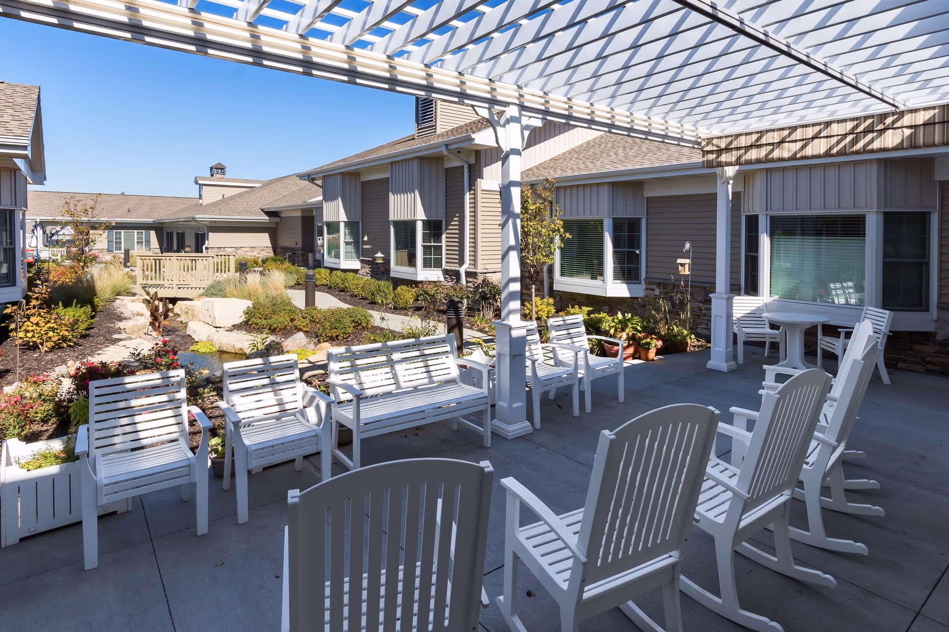 Sunny courtyard with a white pergola and rows of white chairs facing a landscaped garden and surrounding residence buildings.