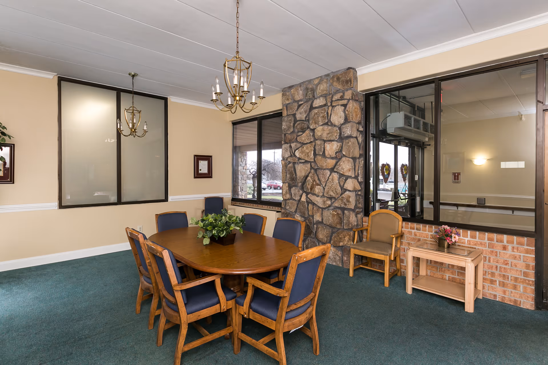 A small dining area with a wooden oval table surrounded by six wooden chairs with blue cushions. The room has green carpet, beige walls, and two chandeliers hanging from the ceiling. There is a stone pillar in the corner and large windows showing an exterior view. A small wooden side table with a flower arrangement and an additional chair are placed near the windows.
