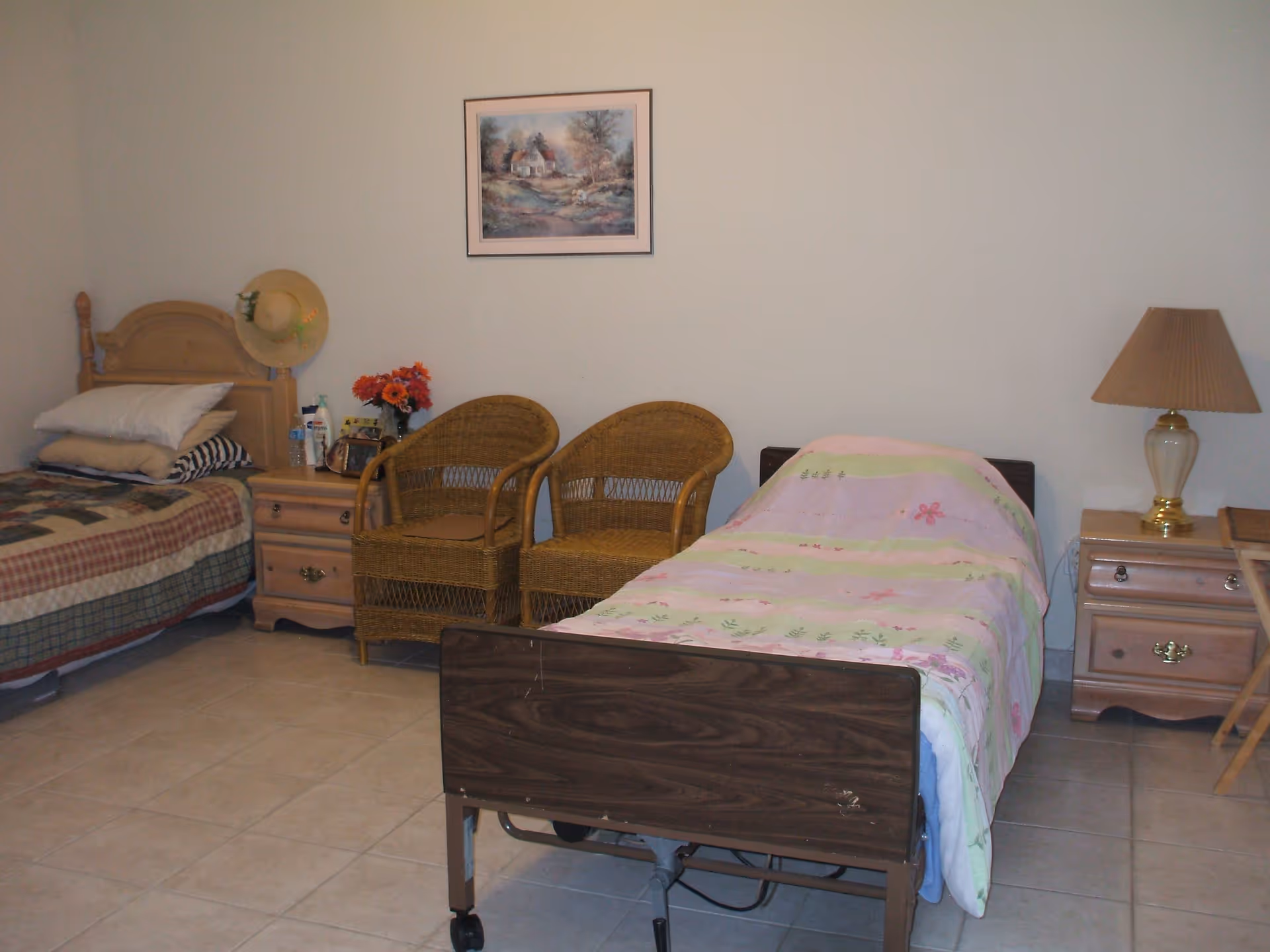 A simple bedroom with two beds, one with a floral pink and green blanket and the other with a plaid blanket. Between the beds are two wicker chairs and a nightstand with a vase of flowers, water bottle, and other small items. Another nightstand with a lamp is beside the bed with the floral blanket. A framed painting hangs on the wall above the chairs.