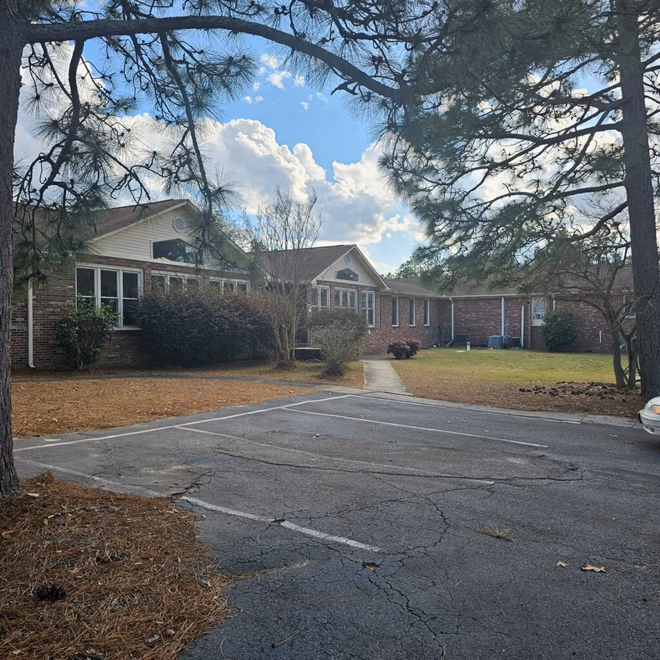 Brick senior living building with windows, shrubs, a walkway, and pine trees framing the entrance