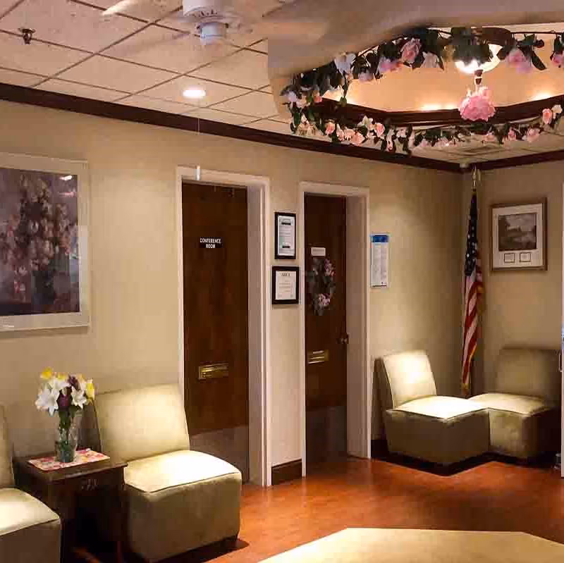 A waiting area with beige cushioned chairs arranged along the walls, a small table with a vase of flowers, two wooden doors labeled 'Conference Room' and another with a wreath, an American flag in the corner, floral artwork on the wall, and a ceiling light fixture decorated with pink flowers.
