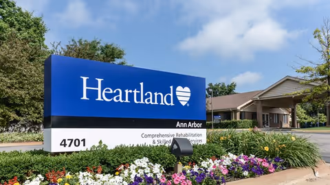 Outdoor view of the entrance sign for Heartland Ann Arbor, a comprehensive rehabilitation and skilled nursing services facility, surrounded by colorful flowers and greenery with a clear blue sky in the background.