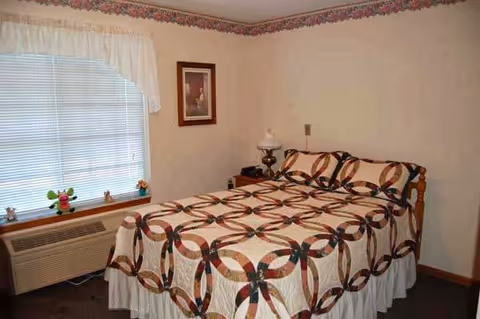 A bedroom with a double bed covered in a quilt with a circular pattern. There is a window with blinds and a white valance on the left side, a framed picture on the wall above the bed, a nightstand with a lamp and a phone, and an air conditioning unit below the window.