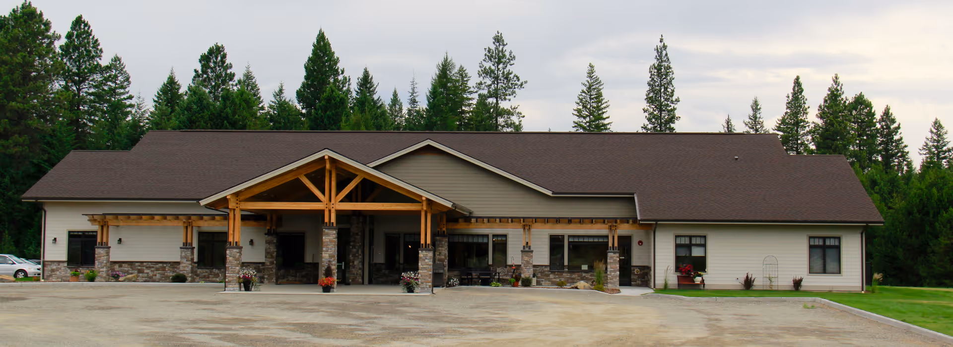 Front exterior view of a single-story assisted living facility building with a large covered entrance supported by wooden beams and stone pillars, surrounded by trees and a gravel driveway.