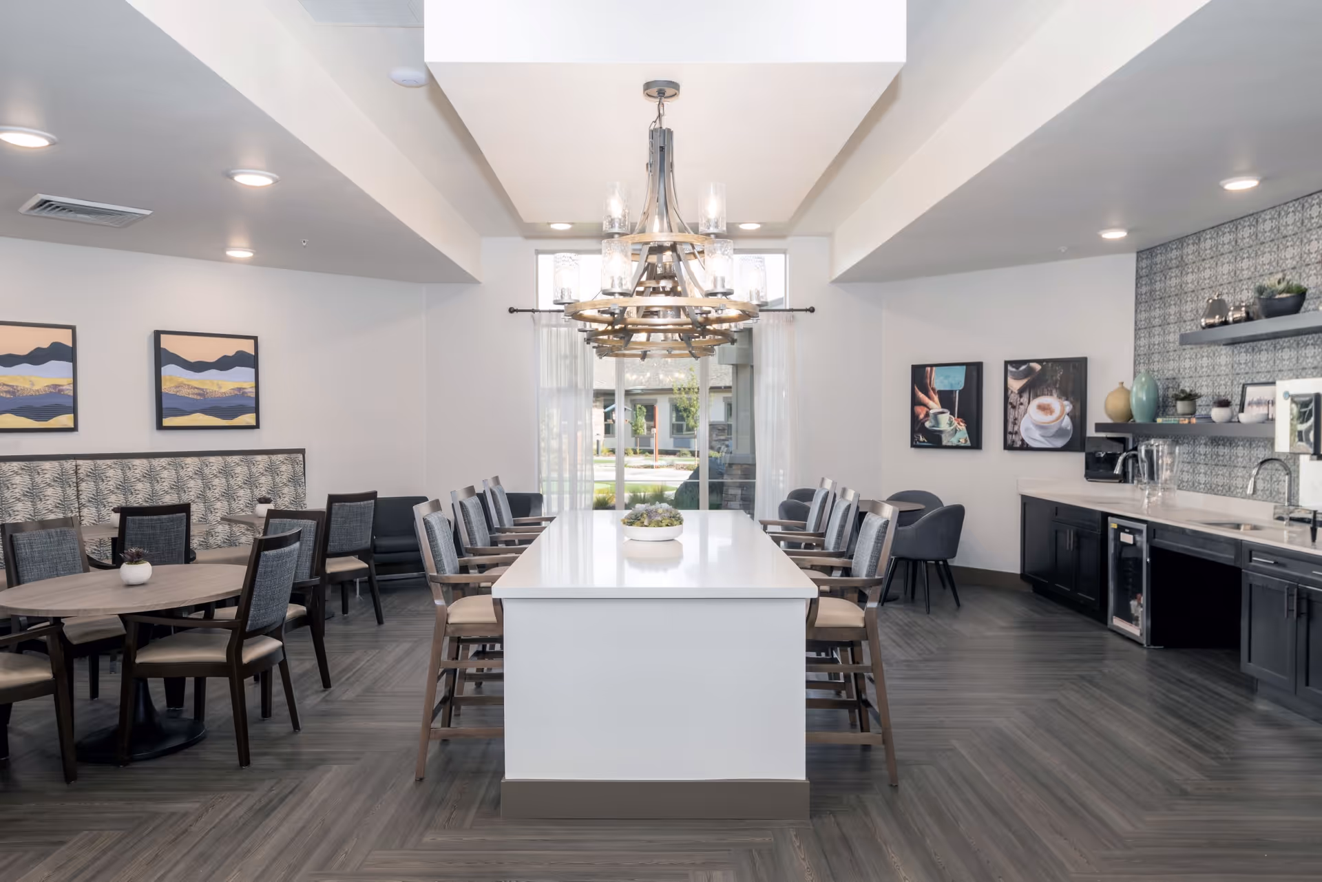 A modern dining room with a long white rectangular table surrounded by wooden chairs with cushions. To the left, there is a round table with chairs and a cushioned bench against the wall. The room has large windows with sheer curtains letting in natural light. On the right side, there is a kitchenette area with a sink, countertop, shelves with decorative items, and framed pictures on the wall. The floor has a dark wood pattern, and a large chandelier hangs above the central table.