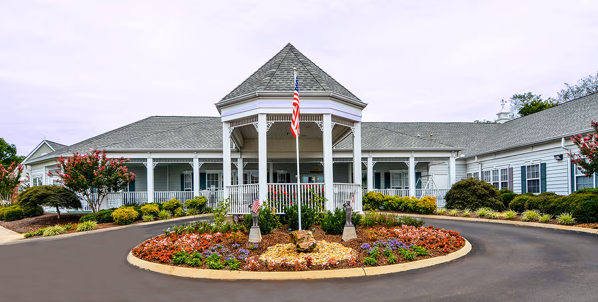 Front exterior view of The Neighborhood at Sheffield facility featuring a circular driveway with a landscaped roundabout in the center, an American flag on a pole, and a white building with a covered porch and decorative trim.