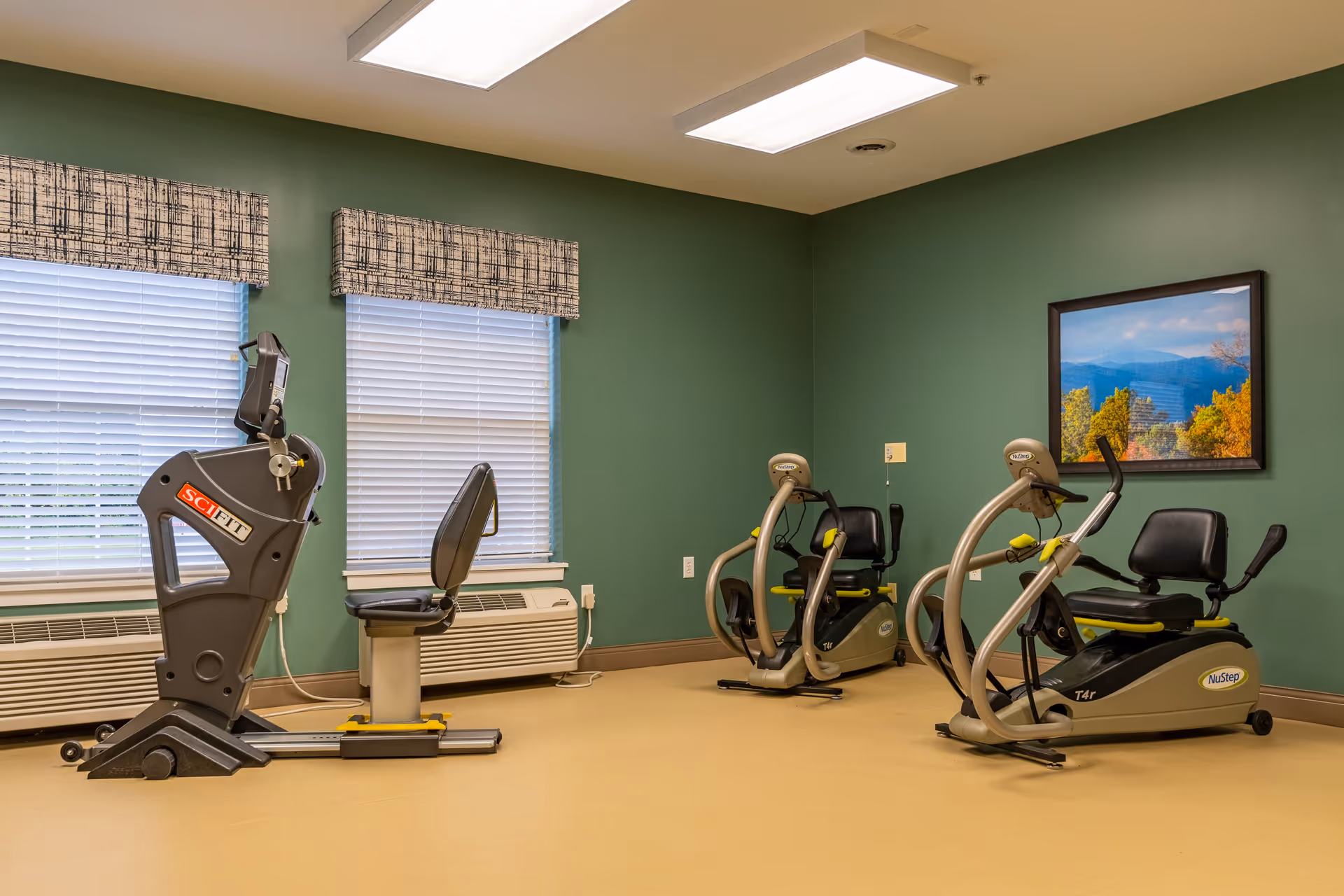 A small exercise room with three recumbent exercise bikes positioned near two windows with blinds and patterned valances. The walls are painted green and there is a framed landscape picture hanging on the wall. The floor is a light tan color and the room is lit by overhead fluorescent lights.