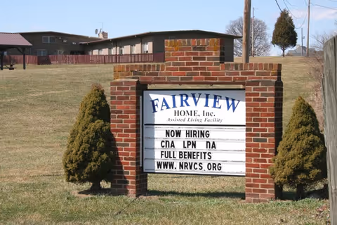 A brick sign structure on a grassy area with two small evergreen trees on either side. The sign reads 'FAIRVIEW HOME, Inc. Assisted Living Facility NOW HIRING CNA LPN NA FULL BENEFITS WWW.NRVCS.ORG'. In the background, there is a building and a covered picnic area.
