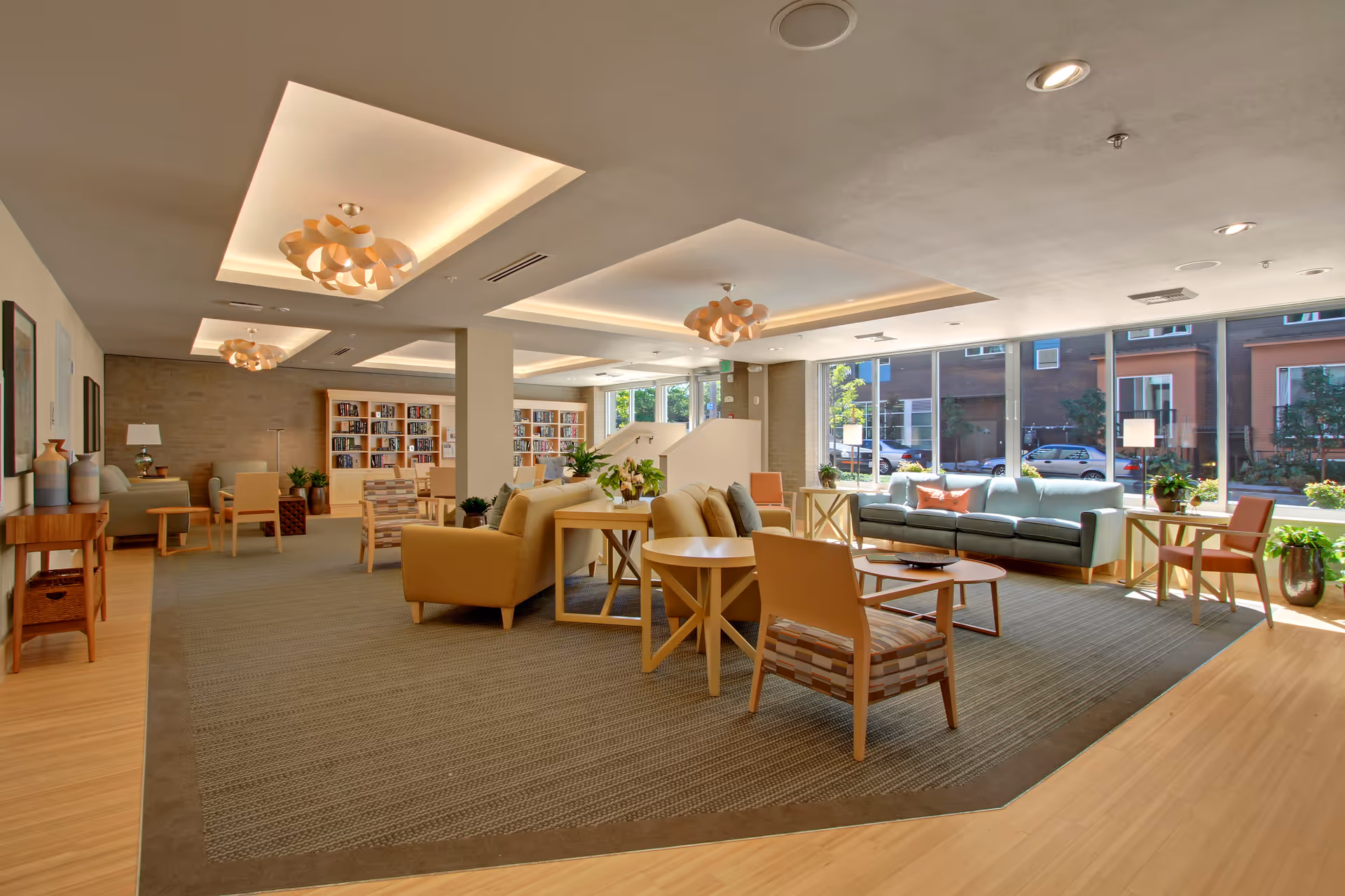 A spacious and well-lit common area in a senior living facility featuring comfortable seating including sofas and armchairs arranged around coffee tables. Large windows allow natural light to fill the room, and bookshelves line the back wall. Modern ceiling lights and indoor plants add to the inviting atmosphere.