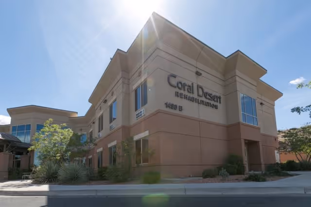 Exterior view of the Coral Desert Rehabilitation And Care building under a clear blue sky with sunlight shining from above. The building is two stories tall with large windows and desert landscaping around it.