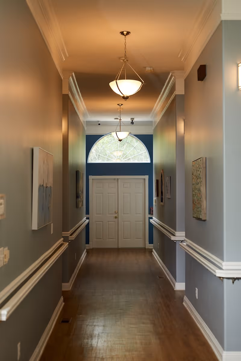 A long, narrow hallway with wooden flooring and light gray walls, decorated with framed artwork on both sides. The hallway is illuminated by ceiling pendant lights and ends with a set of white double doors beneath a large arched window.