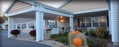 Exterior view of the entrance to Heritage Senior Living At Oakwood Hills, featuring a covered drop-off area with white pillars, seating on the porch, and fall decorations including pumpkins and hay bales near the walkway.