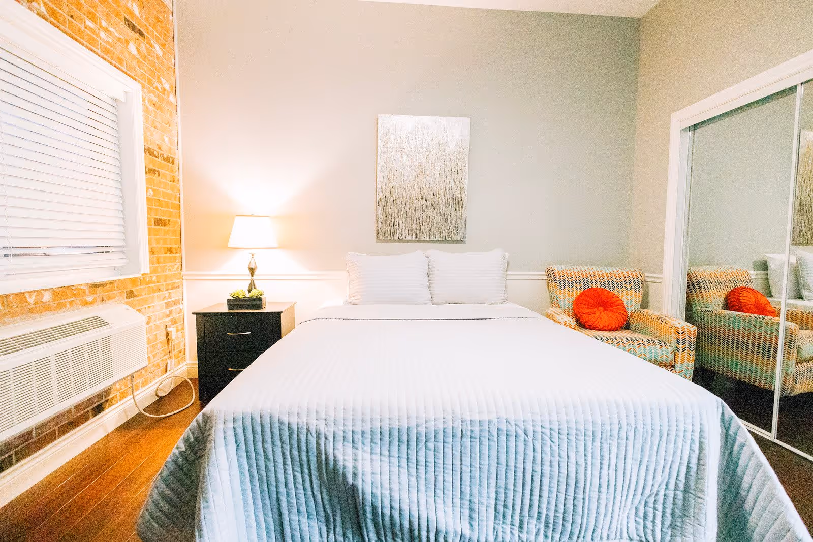 A cozy bedroom with a neatly made bed covered in a white quilt and two white pillows. To the left of the bed is a black nightstand with a lamp and a small plant. The wall behind the bed is painted light gray with a textured artwork hanging above the headboard. To the right is a patterned armchair with an orange round pillow, next to a mirrored sliding closet door. The left wall features exposed brick and a window with white blinds above an air conditioning unit. The floor is wooden.