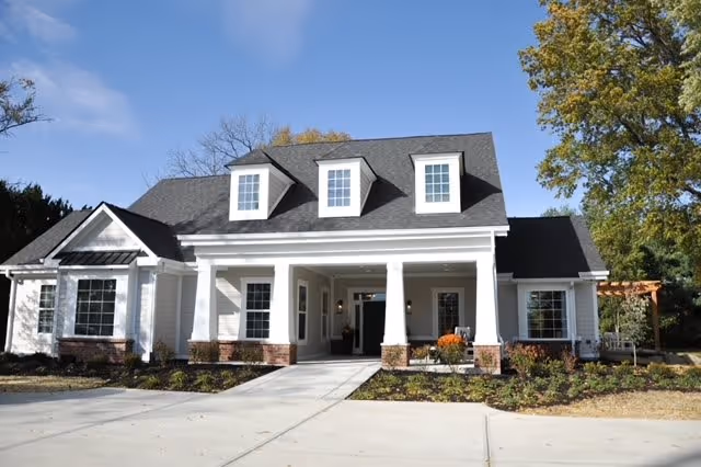 Front exterior view of a single-story building with white siding, brick accents, and a dark gray roof featuring three dormer windows. The entrance has a covered porch supported by white columns, with a concrete driveway and landscaped garden beds in front. Trees and a clear blue sky are visible in the background.
