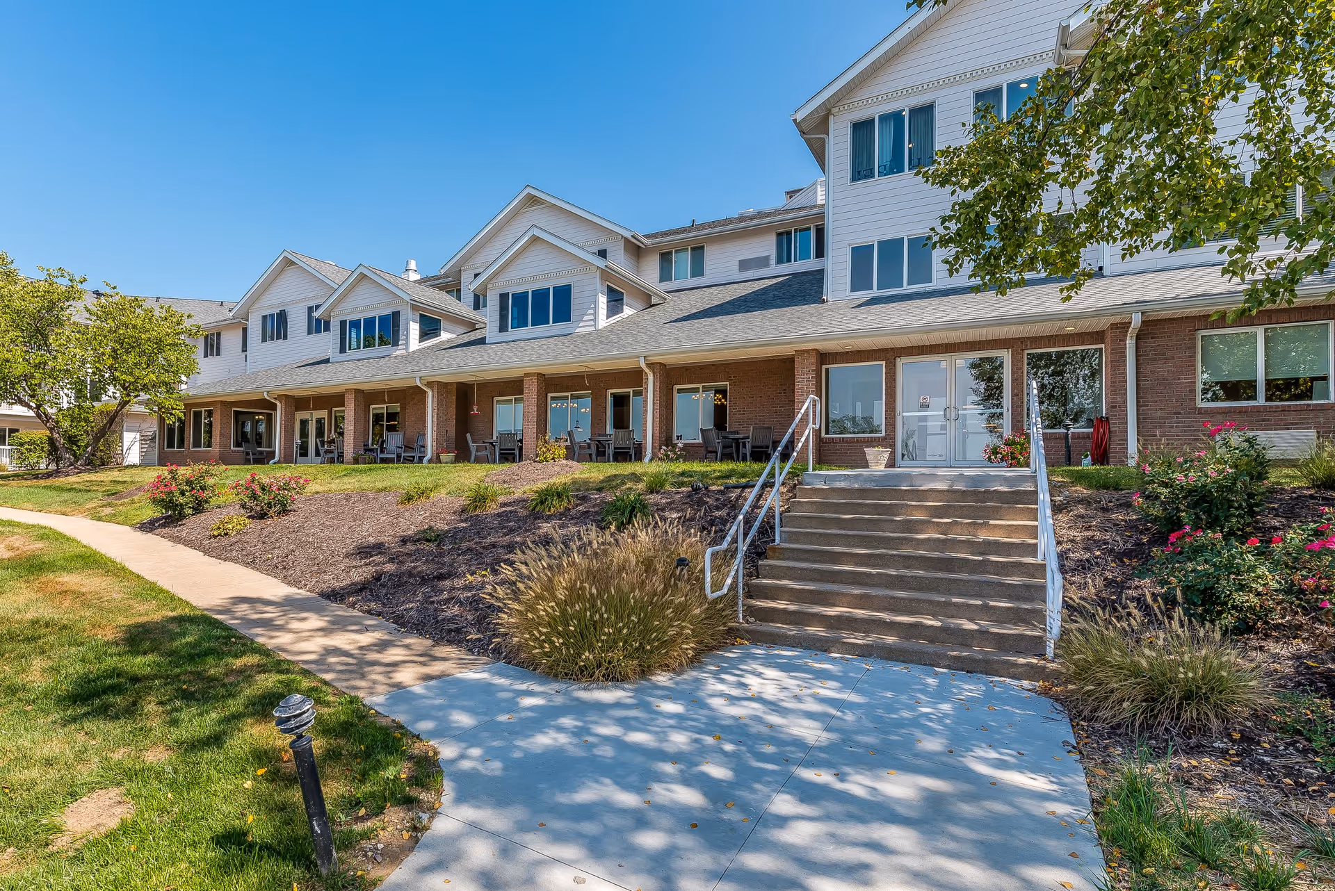 Exterior view of a senior living facility building with a brick and white siding facade, multiple windows, and a covered porch area with chairs. There are steps leading up to double glass doors, surrounded by landscaped bushes and a tree, under a clear blue sky.