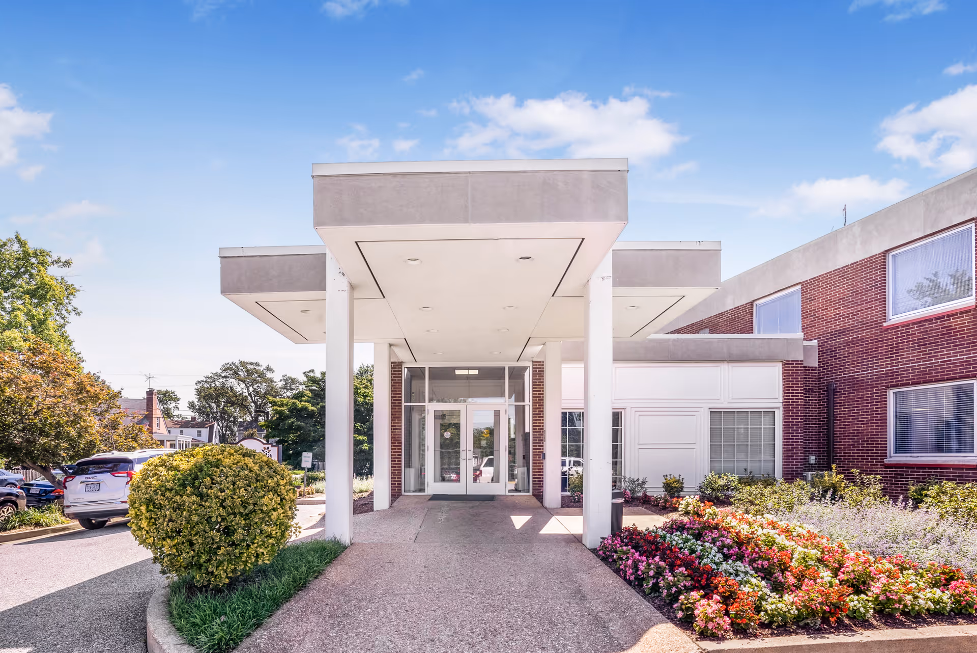 Entrance of Alexandria Rehabilitation & Healthcare Center showing a covered drop-off area with white pillars, a pathway leading to glass double doors, surrounded by well-maintained bushes and colorful flower beds under a clear blue sky.