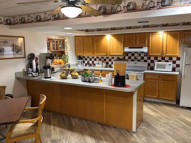 Communal kitchen area with a long serving counter holding coffee and juice dispensers, fruit bowls, and wooden cabinets and appliances.