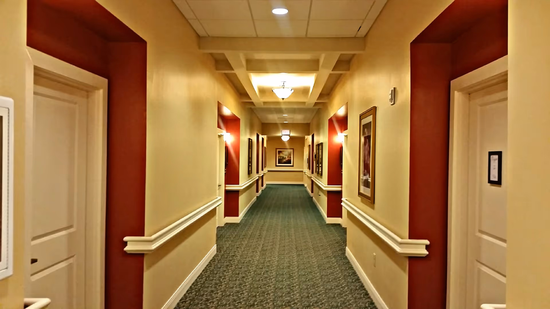 A long, well-lit hallway in a senior living facility with beige walls and red accent panels around the doorways. The carpet is green with a subtle pattern, and framed artwork is hung on the walls. Ceiling lights and wall sconces provide illumination.
