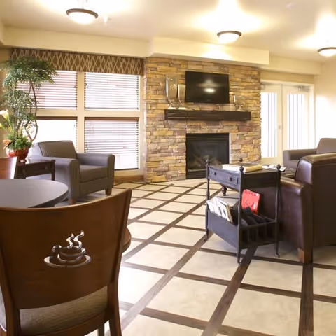 A cozy living room area with a stone fireplace, a mounted flat-screen TV above it, and several armchairs arranged around the room. There is a small table with magazines and books, large windows with blinds, and a potted plant near the window. The floor has a checkered tile pattern and the room is well-lit with ceiling lights.