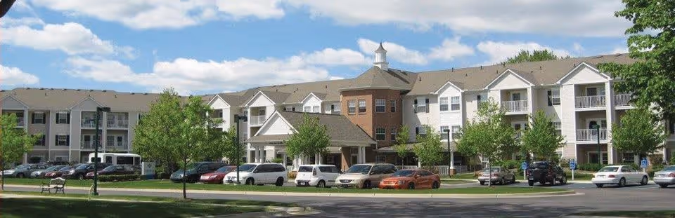 Front exterior of a large three-story senior living building with balconies, landscaped lawn, and parked cars under a partly cloudy sky.