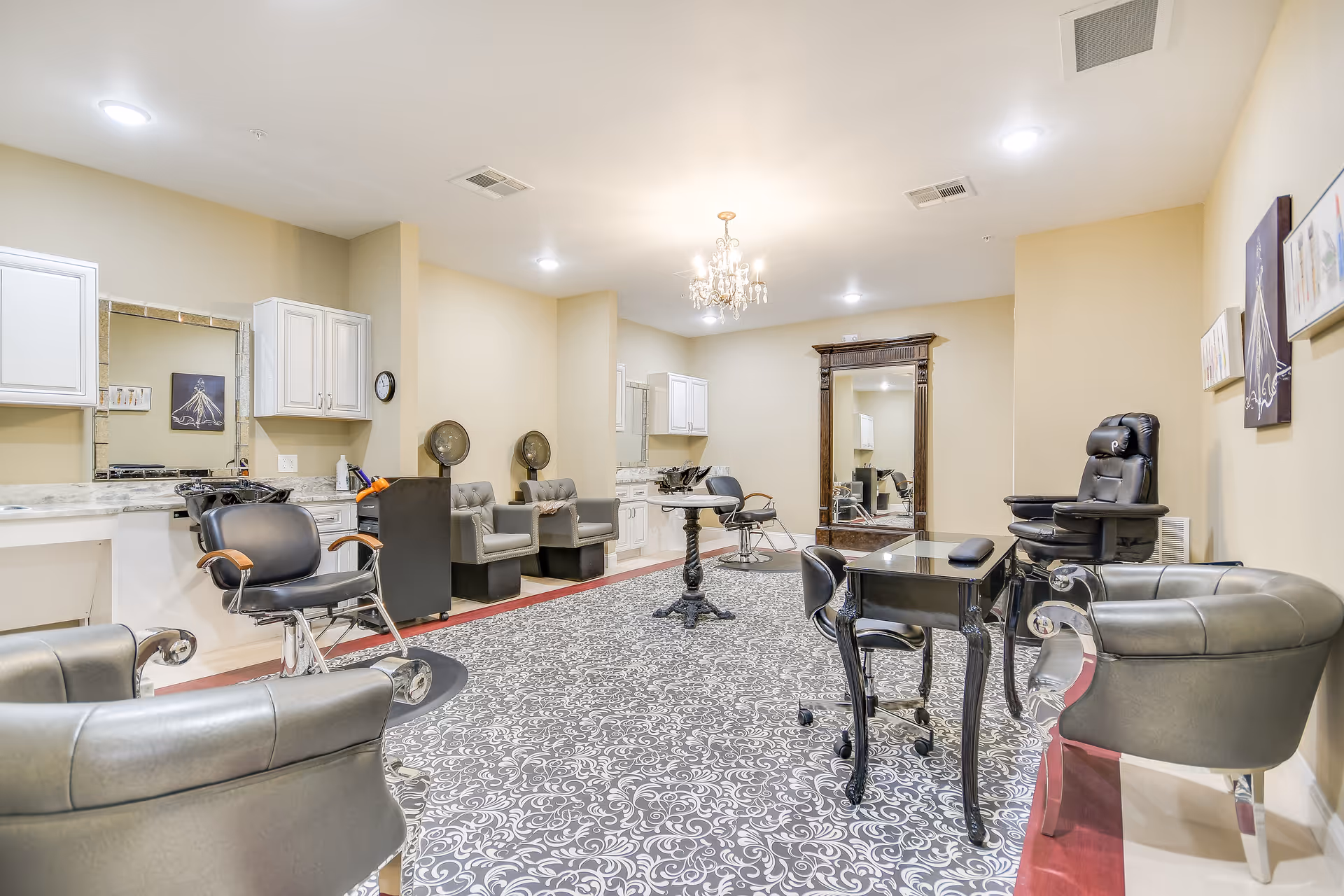 Interior view of a senior living facility's salon area with multiple salon chairs, hair washing stations, a large ornate mirror, and a patterned carpet. The walls are painted beige and there are several pieces of artwork hanging on the walls.