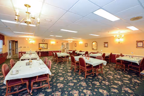 A spacious dining room with multiple tables covered in white tablecloths, each set with cups and condiments. The room has floral carpet flooring, beige walls adorned with framed pictures and mirrors, and is illuminated by chandeliers and wall sconces. There are red cushioned chairs around the tables and a sideboard with a clock and decorative items at the far end.