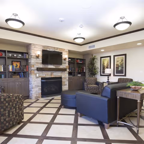 A cozy living room area with a stone fireplace and a mounted flat-screen TV above it. The room features built-in bookshelves on either side of the fireplace filled with books and decorative items. There are several upholstered chairs and a black leather sofa arranged around a central ottoman. The floor has a checkered tile pattern, and the walls are adorned with framed artwork and a potted plant. Ceiling lights provide bright illumination.