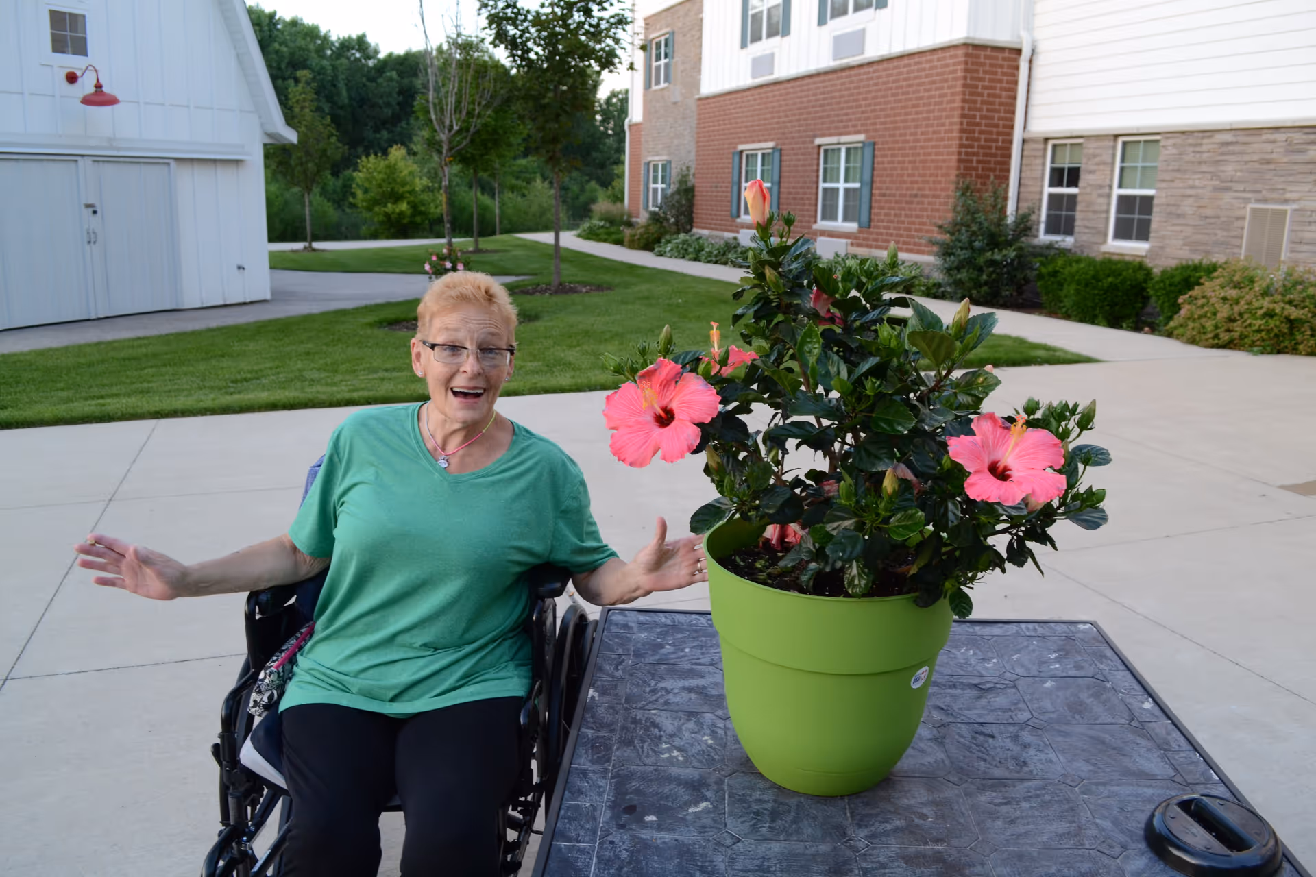 An elderly woman in a wheelchair wearing a green shirt and glasses is smiling and gesturing with her hands outdoors near a table with a large green pot containing pink hibiscus flowers. Behind her is a building with brick and stone exterior and a well-maintained lawn with trees and shrubs.