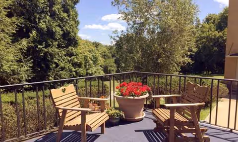 Balcony with two wooden chairs, railing, and red potted flowers overlooking trees.