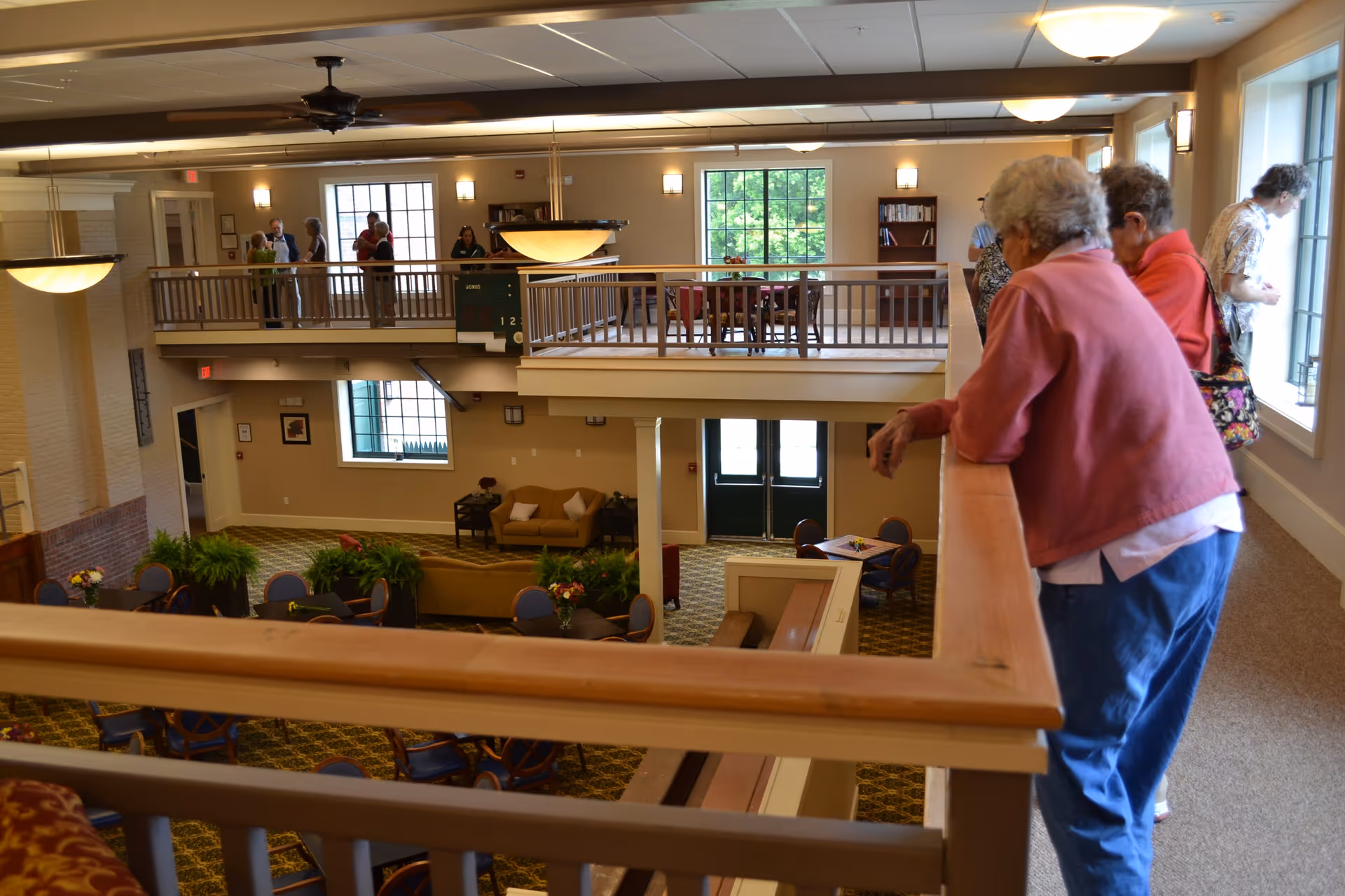 Interior view of a senior living community common area with multiple elderly people standing and looking over a wooden railing. The space includes seating areas with chairs and tables, large windows letting in natural light, and a carpeted floor with plants and flowers placed around the room.