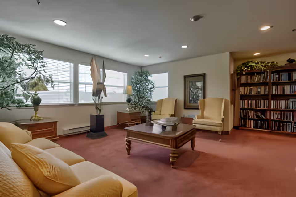 A cozy living room with beige armchairs and a sofa surrounding a wooden coffee table. The room has large windows letting in natural light, a tall plant, a sculpture on a pedestal, a framed picture on the wall, and a bookshelf filled with books.
