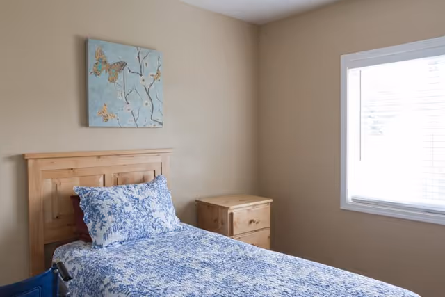 A simple bedroom with a wooden bed frame and a bed made with blue and white patterned bedding. There is a wooden nightstand next to the bed and a window with white blinds letting in natural light. A decorative painting featuring butterflies hangs on the beige wall above the bed.