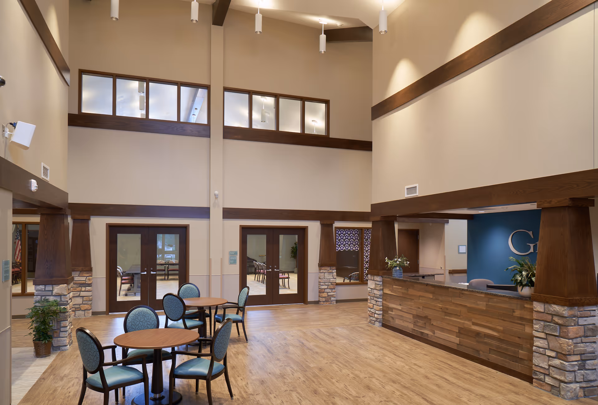 Spacious and well-lit interior common area with high ceilings, featuring a wooden reception desk with stone accents, round tables with blue cushioned chairs, and large windows and doors leading to other rooms.
