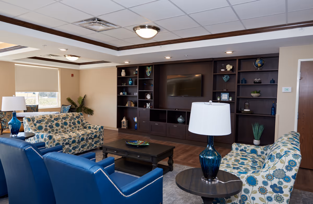 A cozy living room in a senior living facility featuring two floral patterned sofas, two blue armchairs, a dark wooden coffee table, and a matching side table with a blue glass lamp. The room has a large built-in dark wood entertainment center with shelves holding decorative items and a flat-screen TV mounted in the center. There is a window with a beige roller shade and a potted plant in the corner.