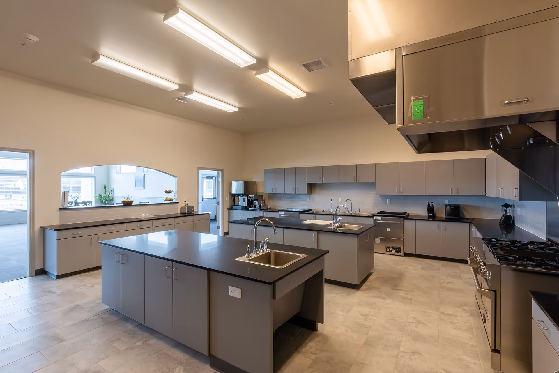 A spacious, modern kitchen with two large islands featuring sinks, gray cabinets, stainless steel appliances including a large stove with an overhead vent, and bright overhead lighting. There is a pass-through window to an adjacent room with plants and fruit bowls visible.